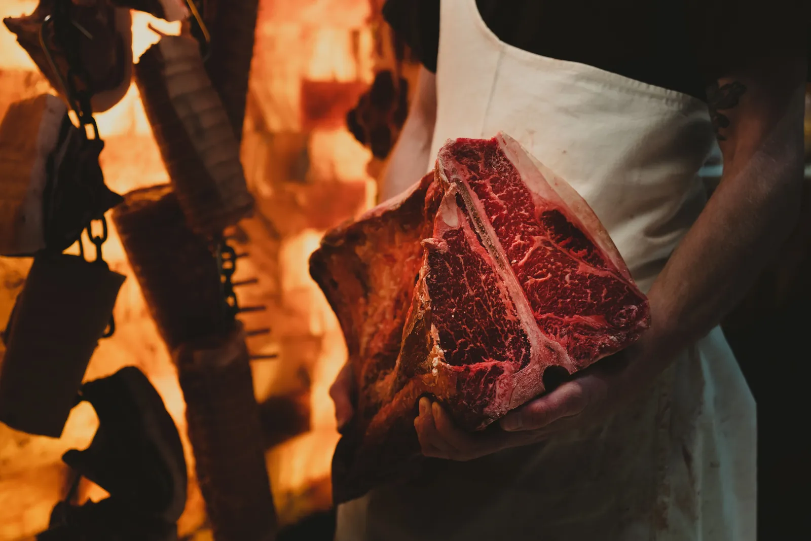 A butcher in a white apron holding a marbled bone-in steak in front of a fire