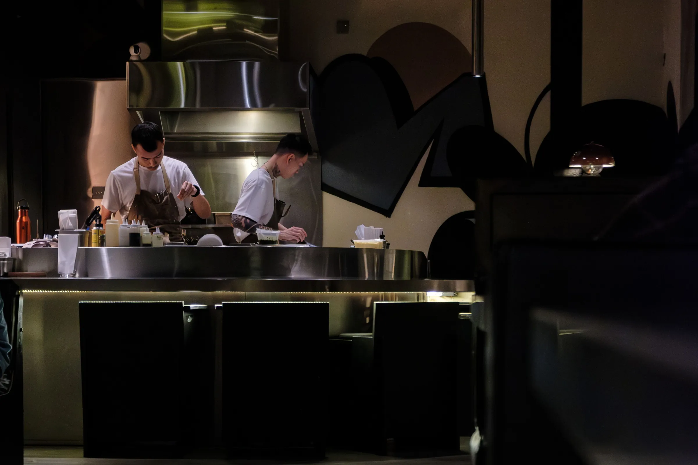 Two chefs at the pass of a dark open kitchen, steel counters gleaming