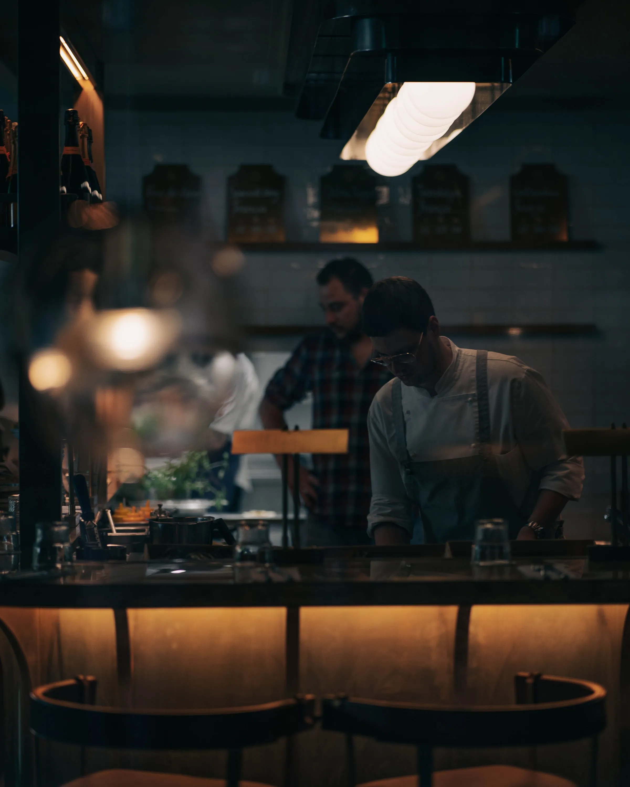 Two chefs working behind the pass in a dim open kitchen, seen from the counter stools