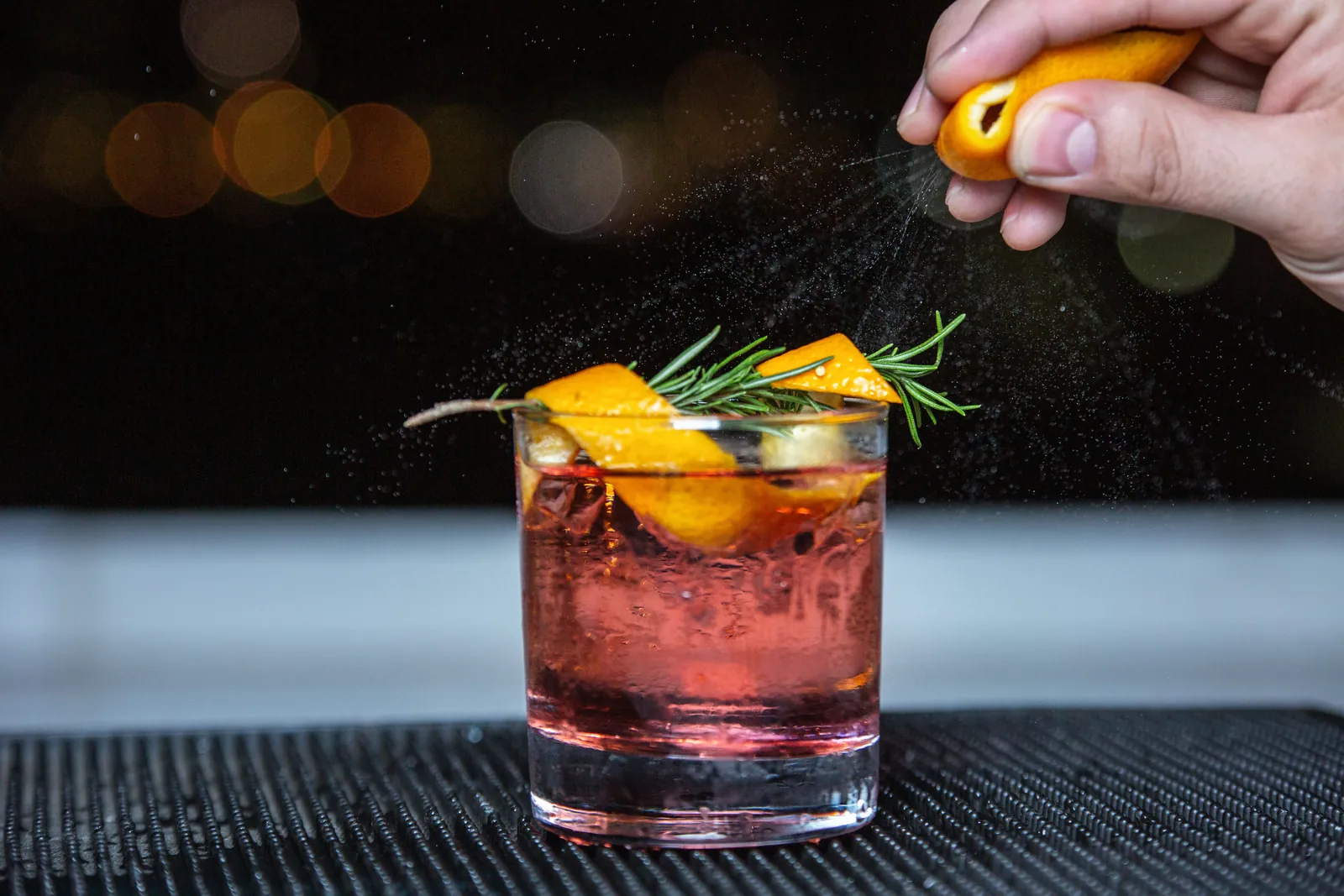 A red cocktail with a sprig of rosemary and a twist of orange being sprayed