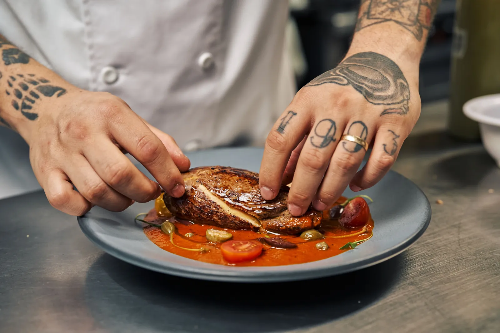A chef&rsquo;s tattooed hands laying a slice of steak onto a plate