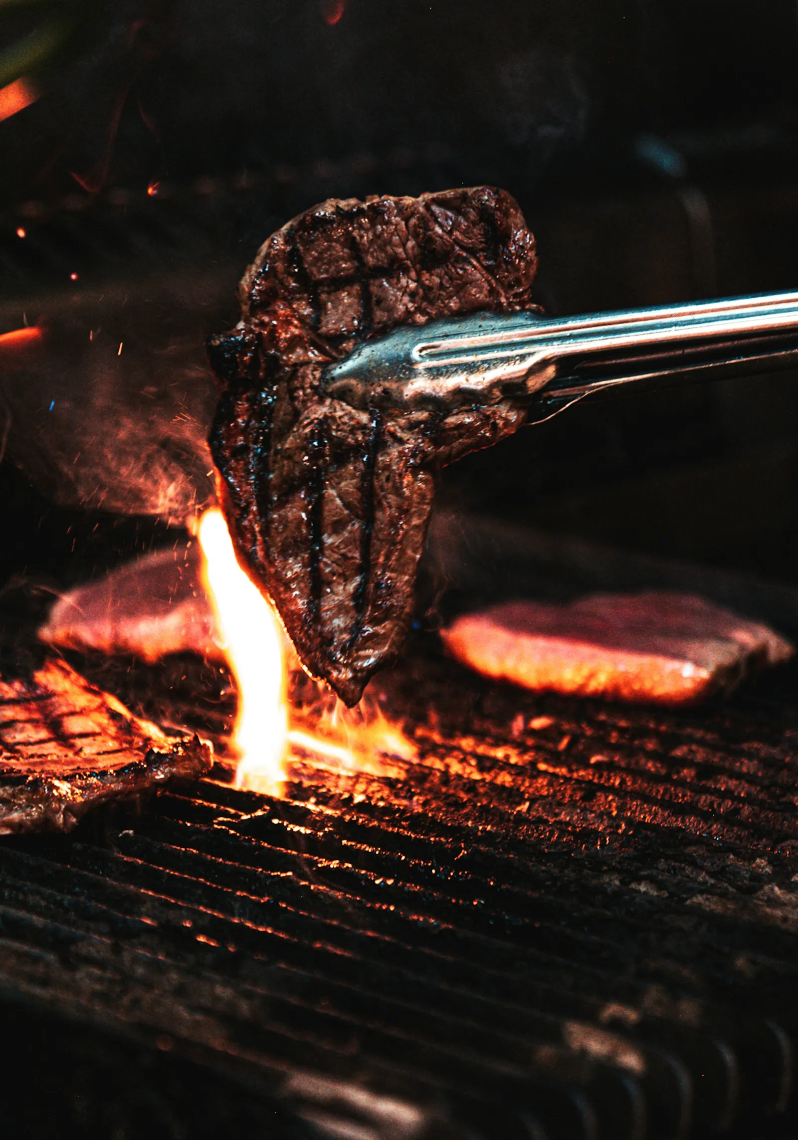 Tongs lifting a thick seared steak off the grill, flames underneath