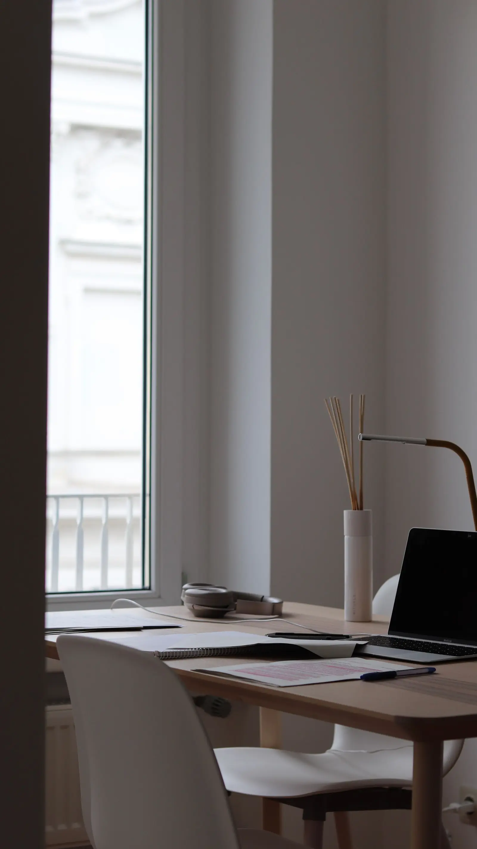 A pale desk and chair set against a tall window in a room with plain walls.