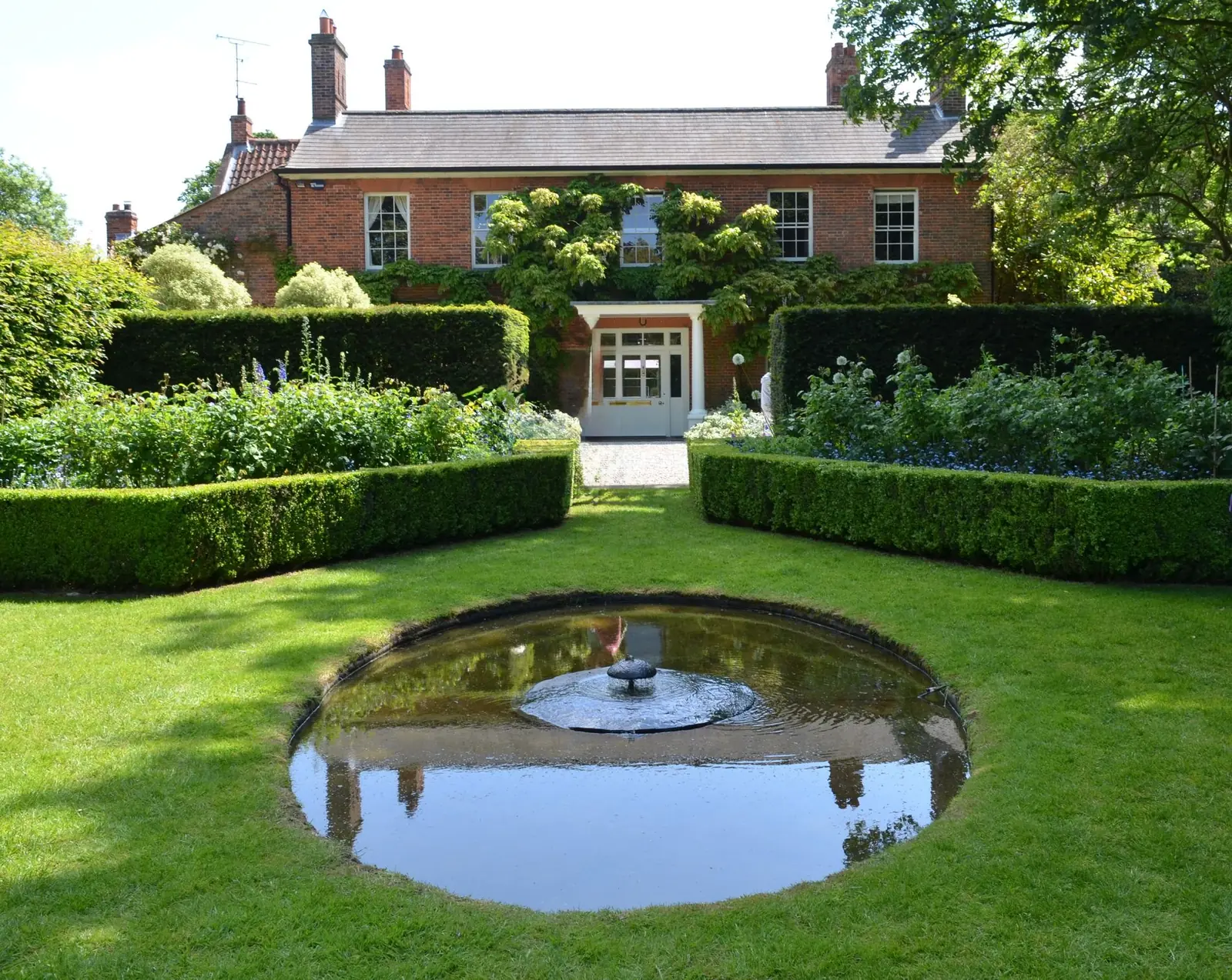 A formal English garden with clipped hedges, a circular reflecting pool and a red-brick house behind.