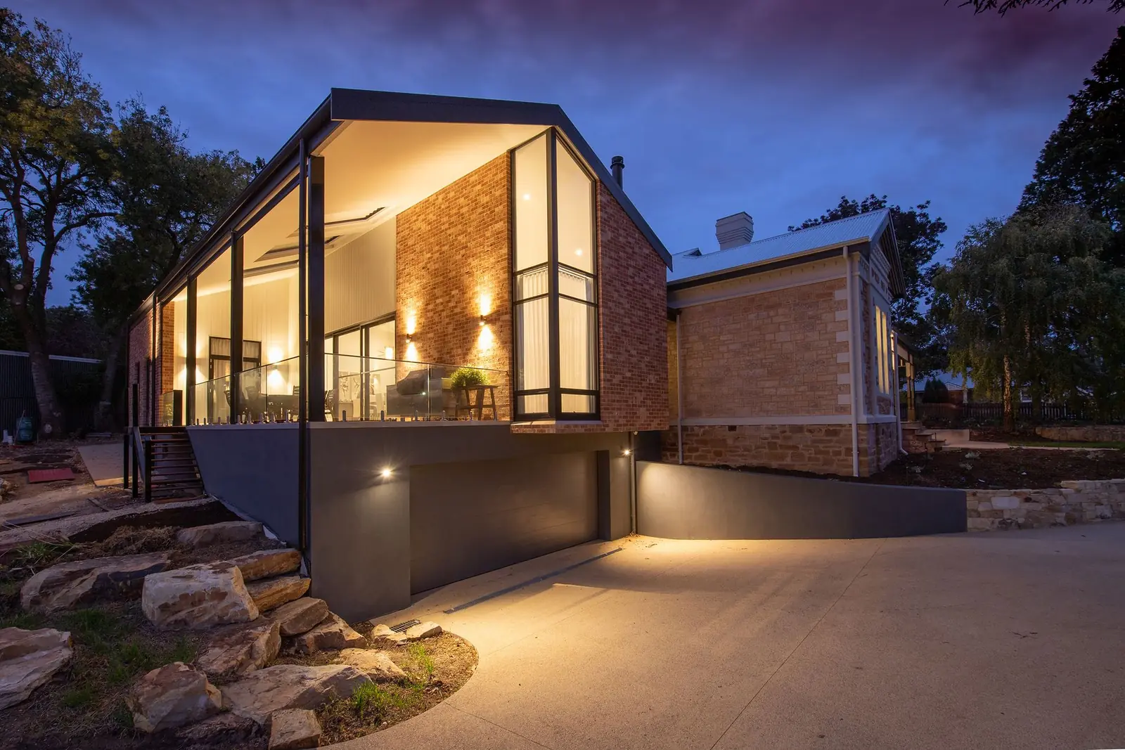 A glass-fronted garden building at dusk, lit from inside, with steps and planting lit below it.