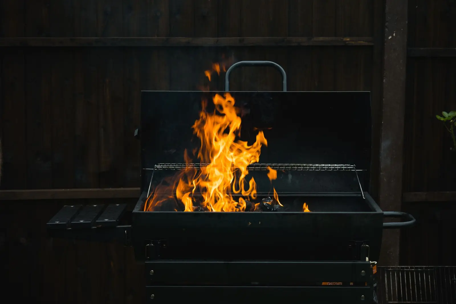 A charcoal barbecue burning against a dark timber garden fence in the evening.