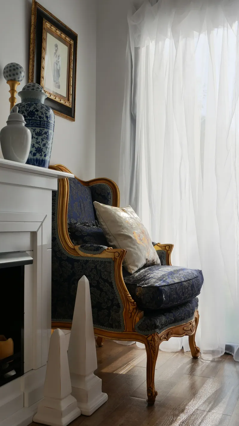 An upholstered armchair beside a painted fireplace, with a cushion and a long curtain.