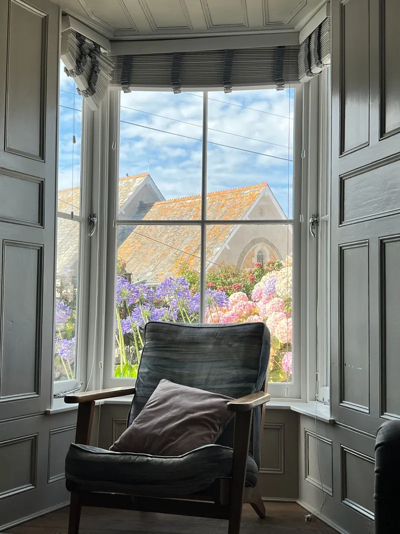 A chair at an open casement window looking out over hydrangeas and a stone roof.